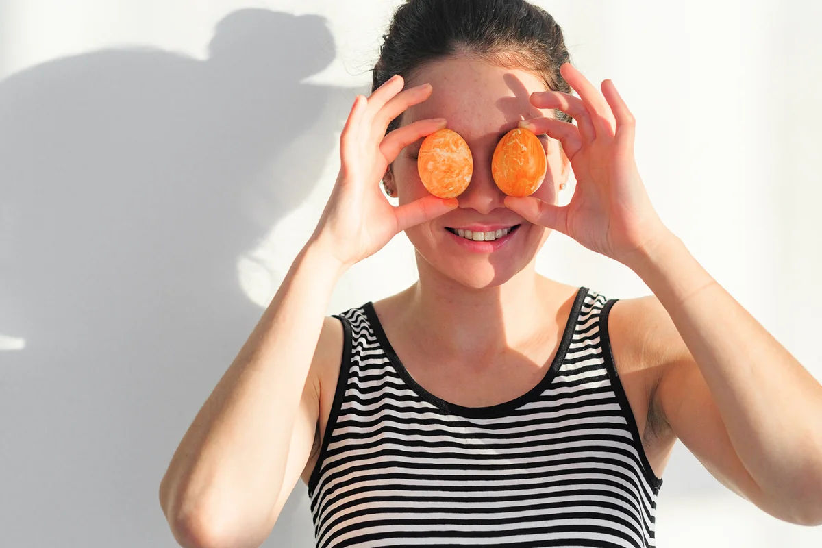 Portrait Of Woman Holding Eggs On Eyes While Standing Against Wall