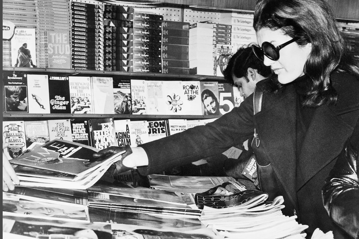 Mrs. Jacqueline Onassis purchases magazines at airport bookshop before boarding flight at Heathrow