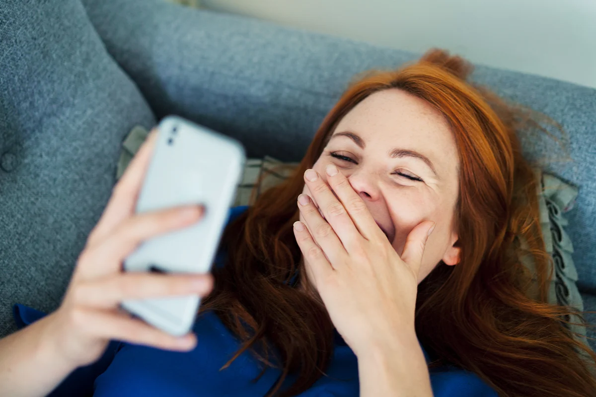 Woman lying on sofa using smart phone, smiling behind hand
