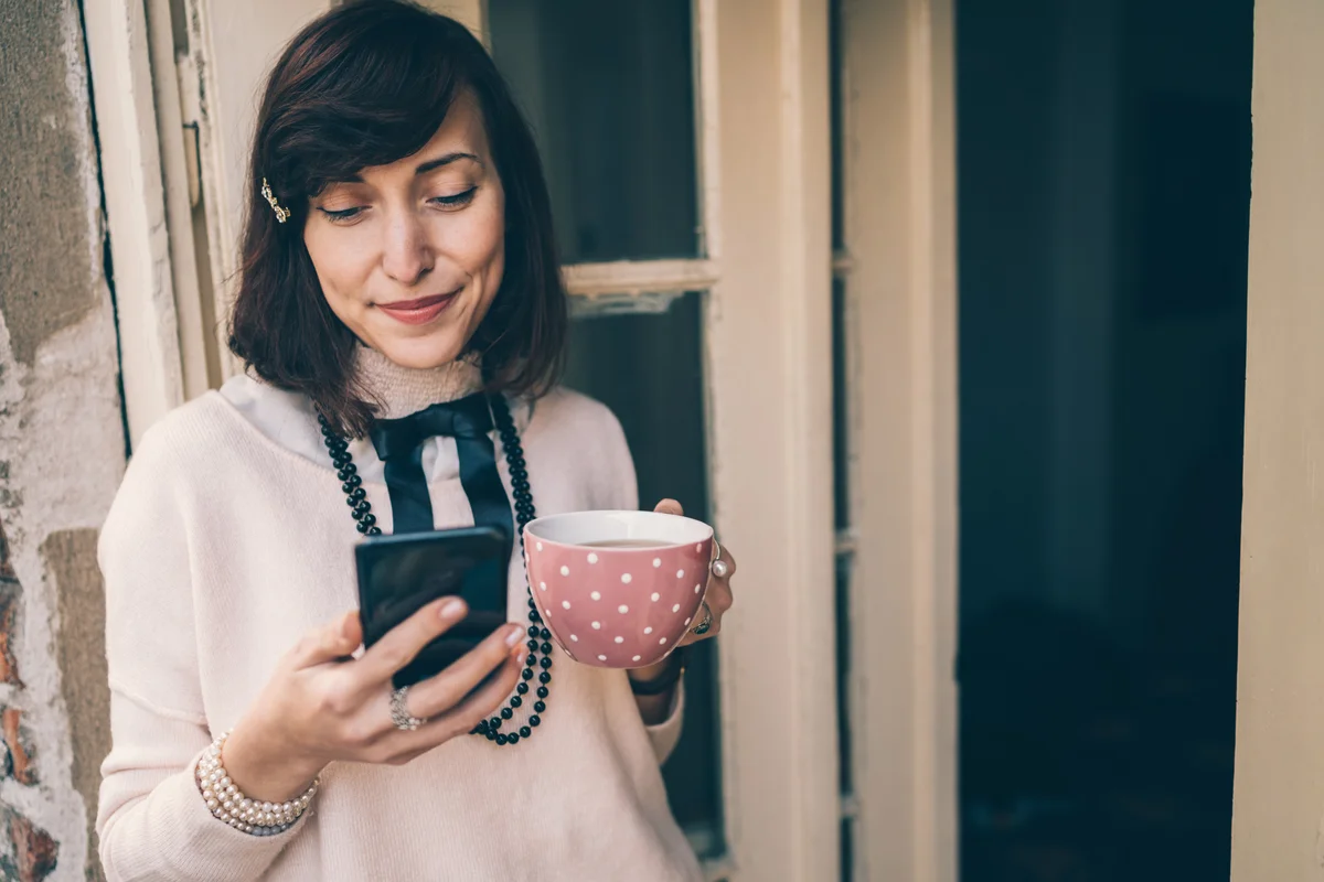 Smiling woman wearing black beads, using smartphone and drinking warm drink