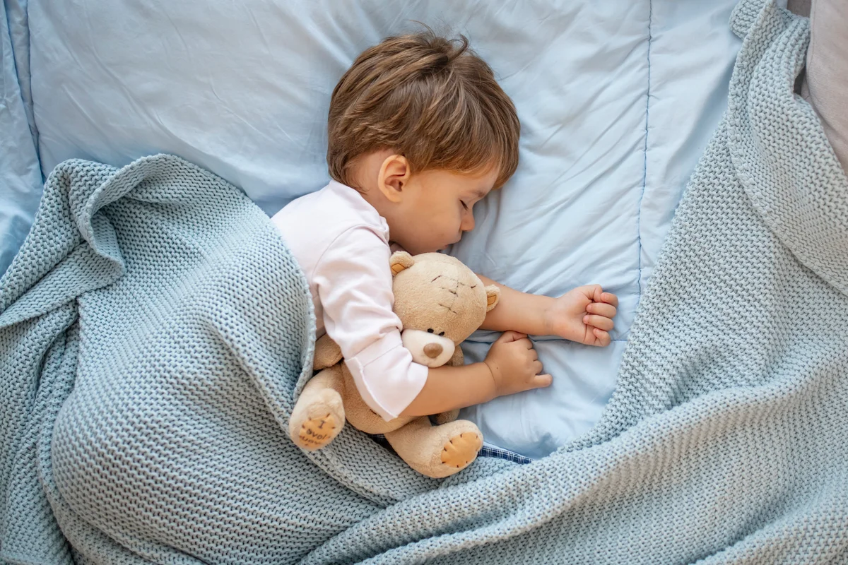 toddler boy sleeping in bed with light blue blanket and teddy bear