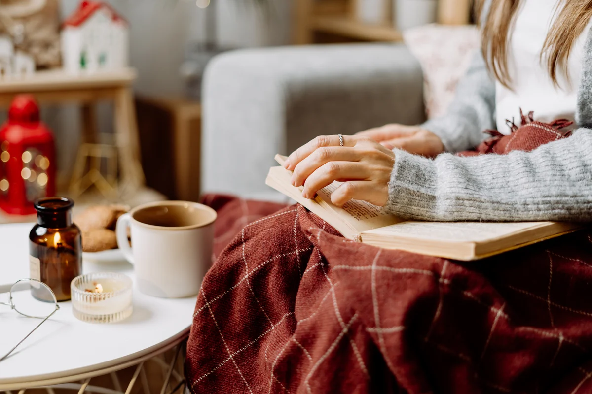 Woman curled up in blanket reading book with hot drink in a mug