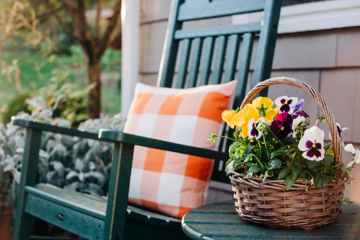 Close-up of a basket filled with pansies next to a green rocking chair with orange pillow
