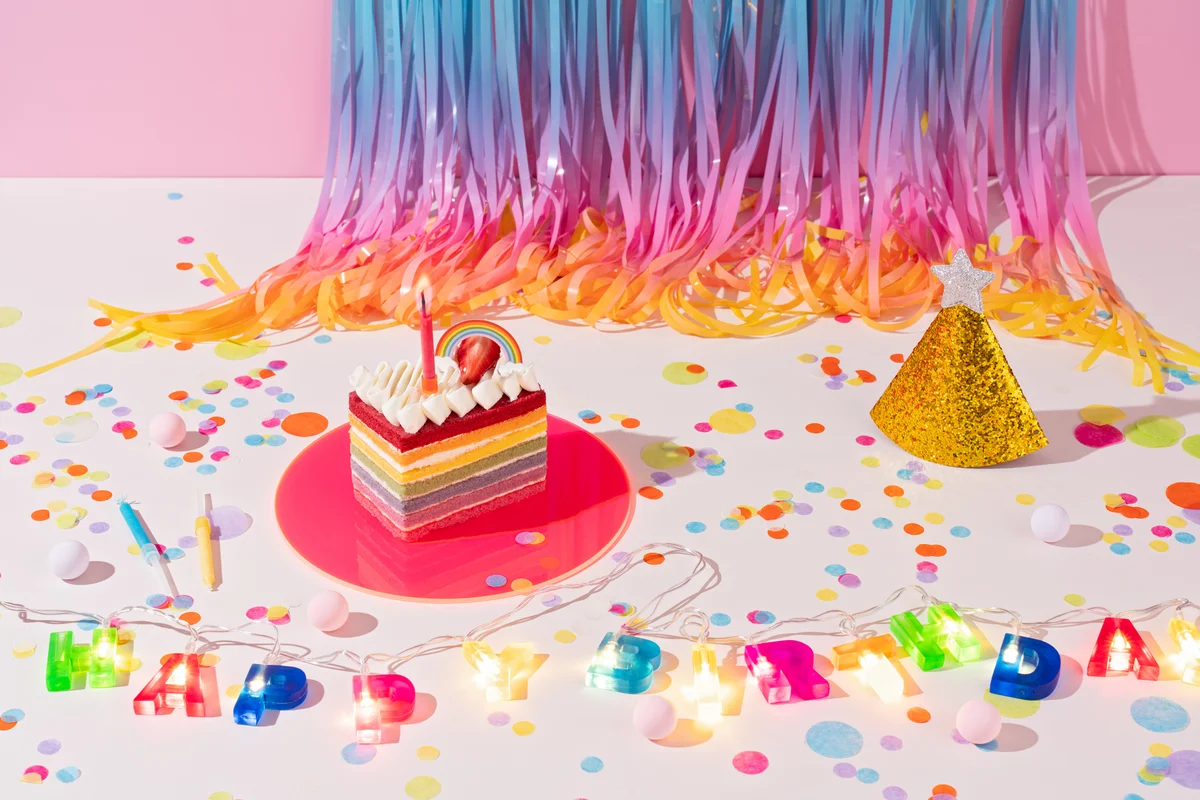 Piece of rainbow cake and a glittery birthday hat, rainbow tinsel in background