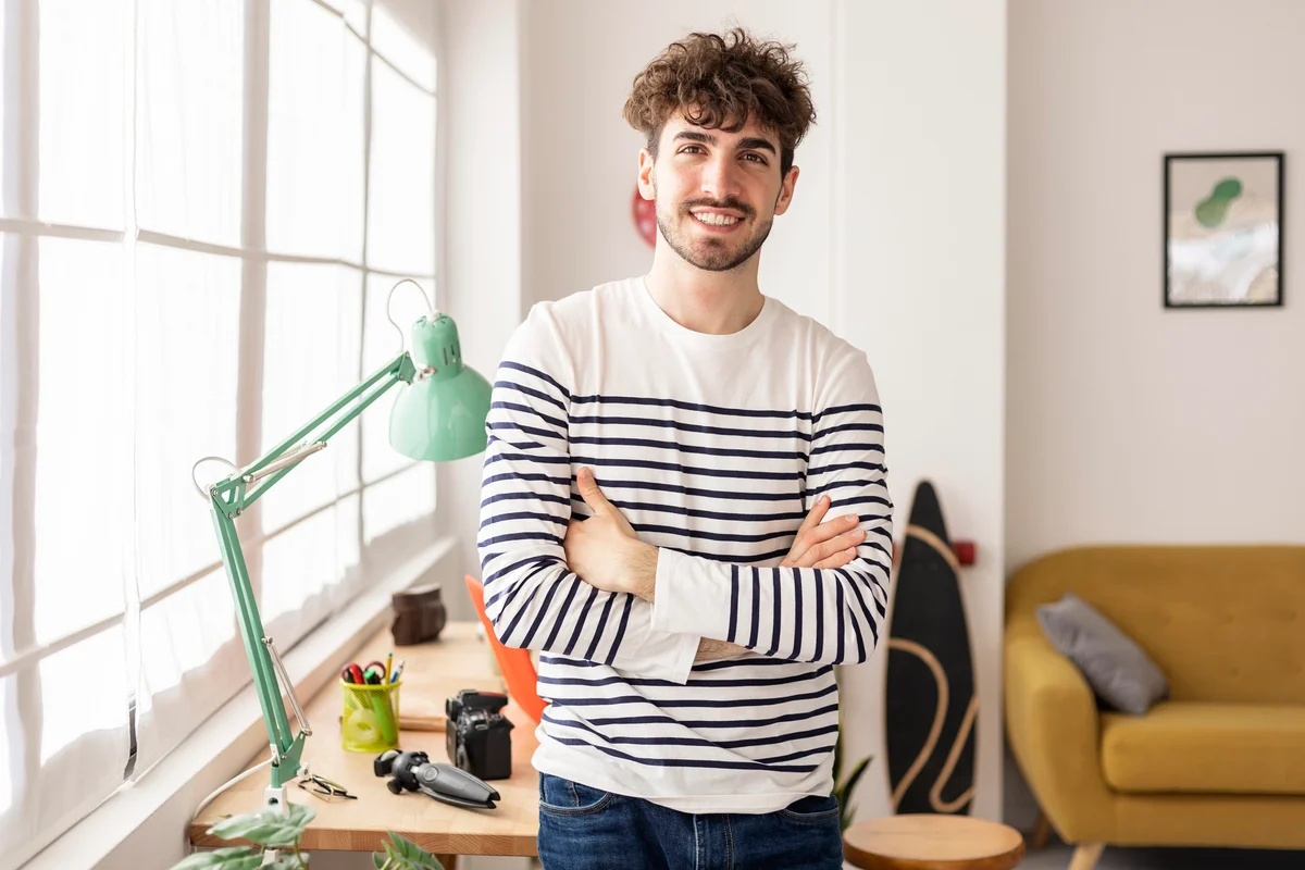 Front view of young man posing with crossed arms in his workspace at home looking at camera