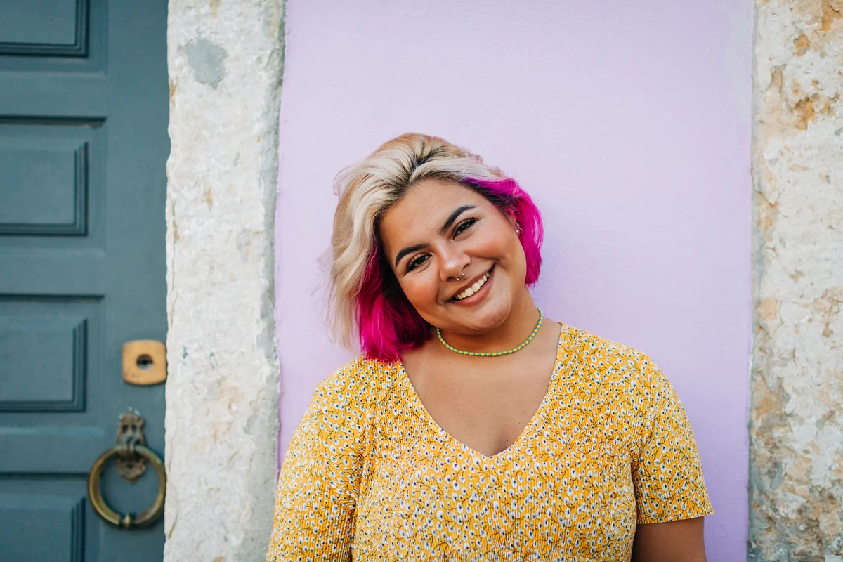 Girl with bright pink hair, yellow shirt in front of purple door and stone walls