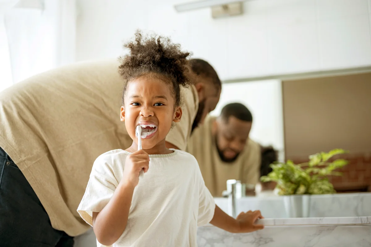 Kid brushing her teeth while her father stands behind in the bathroom