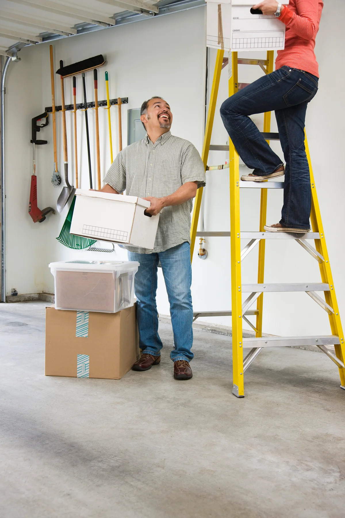 Woman climbing ladder to garage ceiling while man hands her boxes