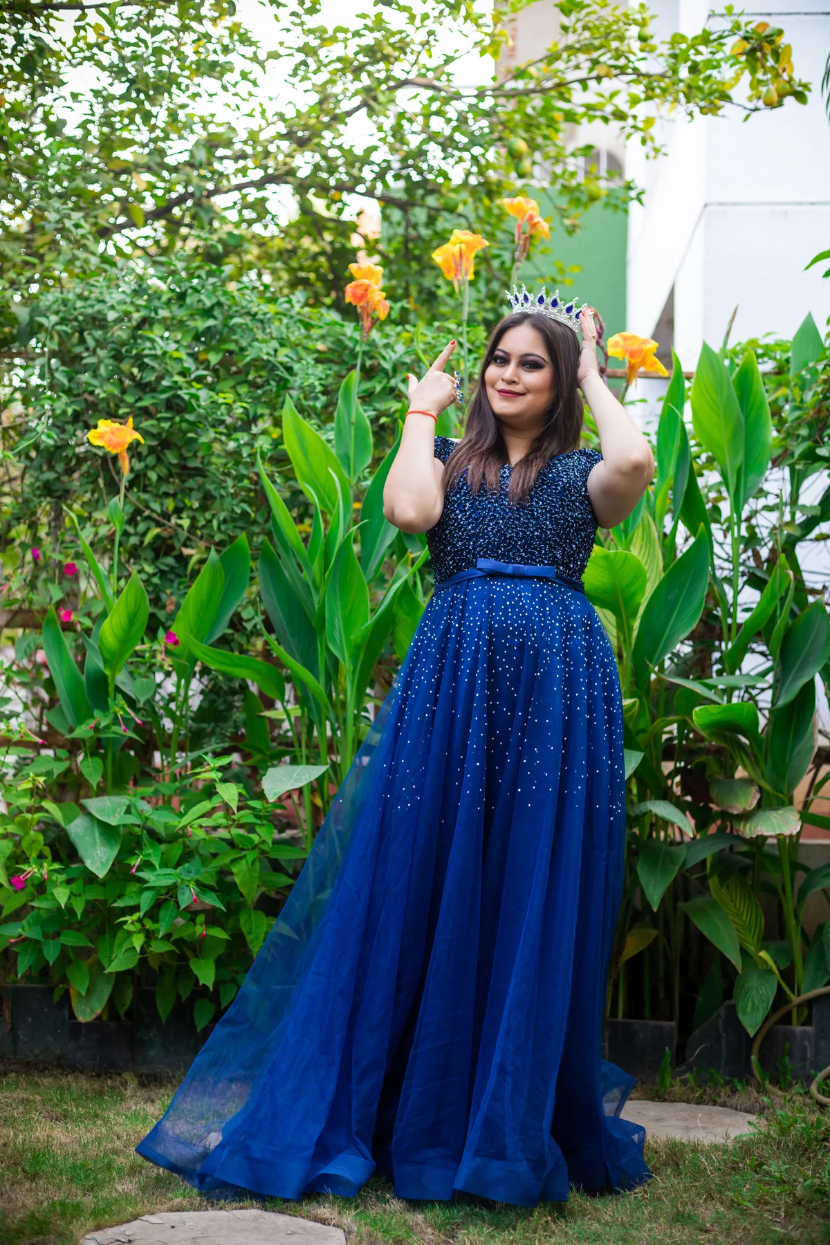 Girl wearing crown and navy dress standing near shady plants and pointing to crown