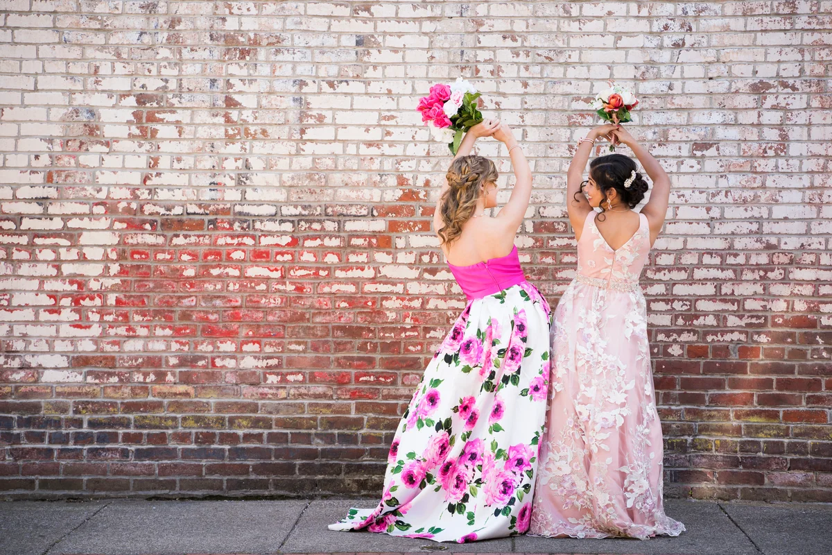 Two girls dancing in prom dresses in front of brick wall