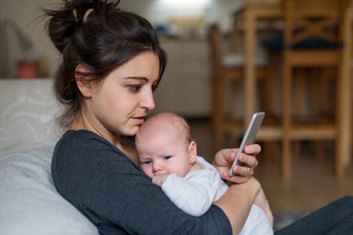 Loving and affectionate mother holding newborn baby indoors at home, using smartphone.