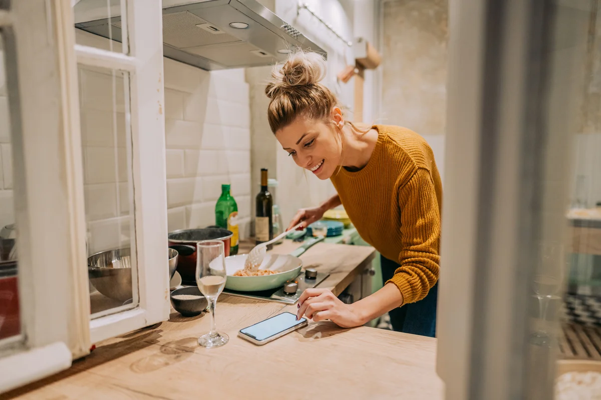 Photo of a young woman cooking homemade pasta and checking recipe online with a glass of wine