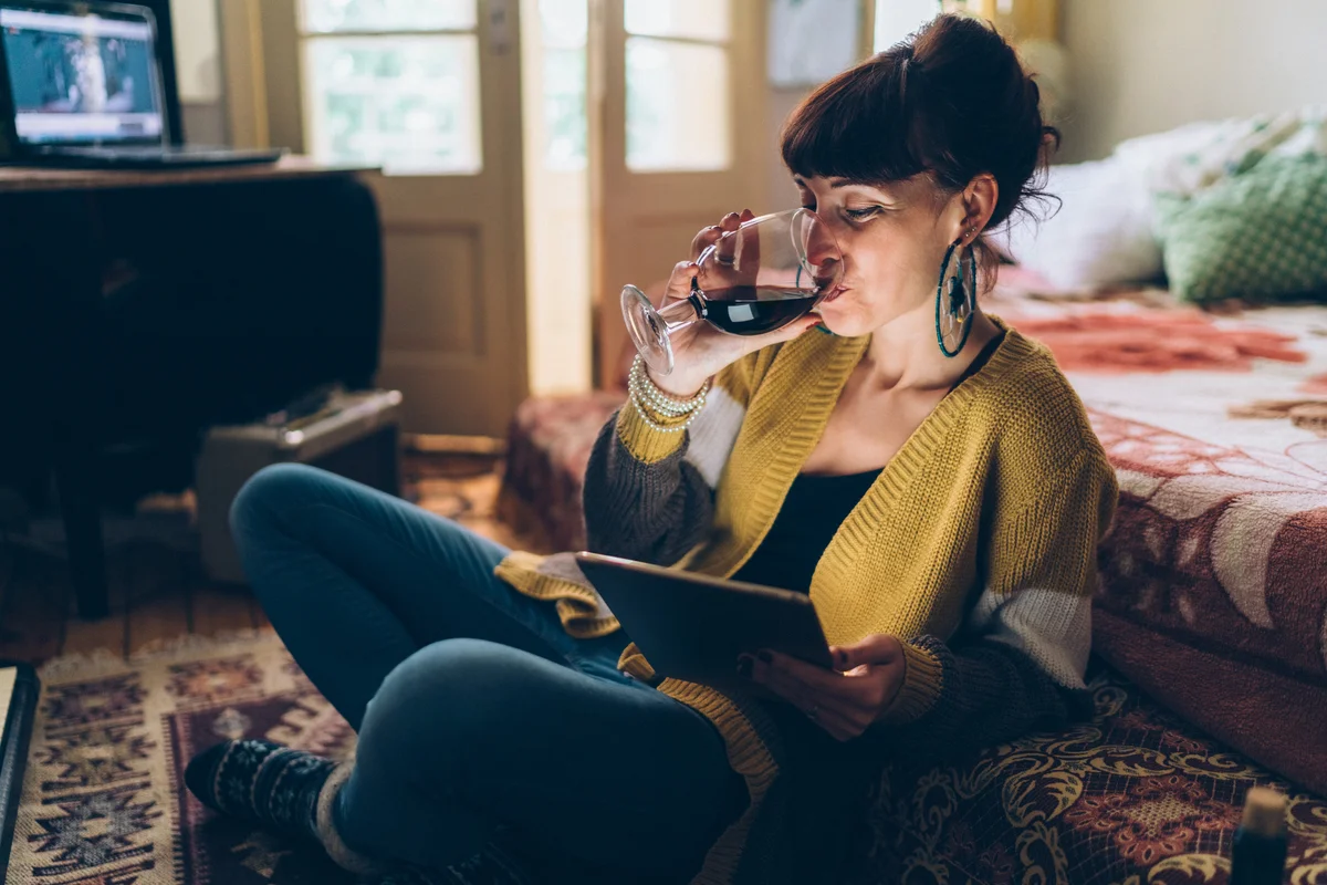 Woman enjoying glass of wine in dark room while on her tablet