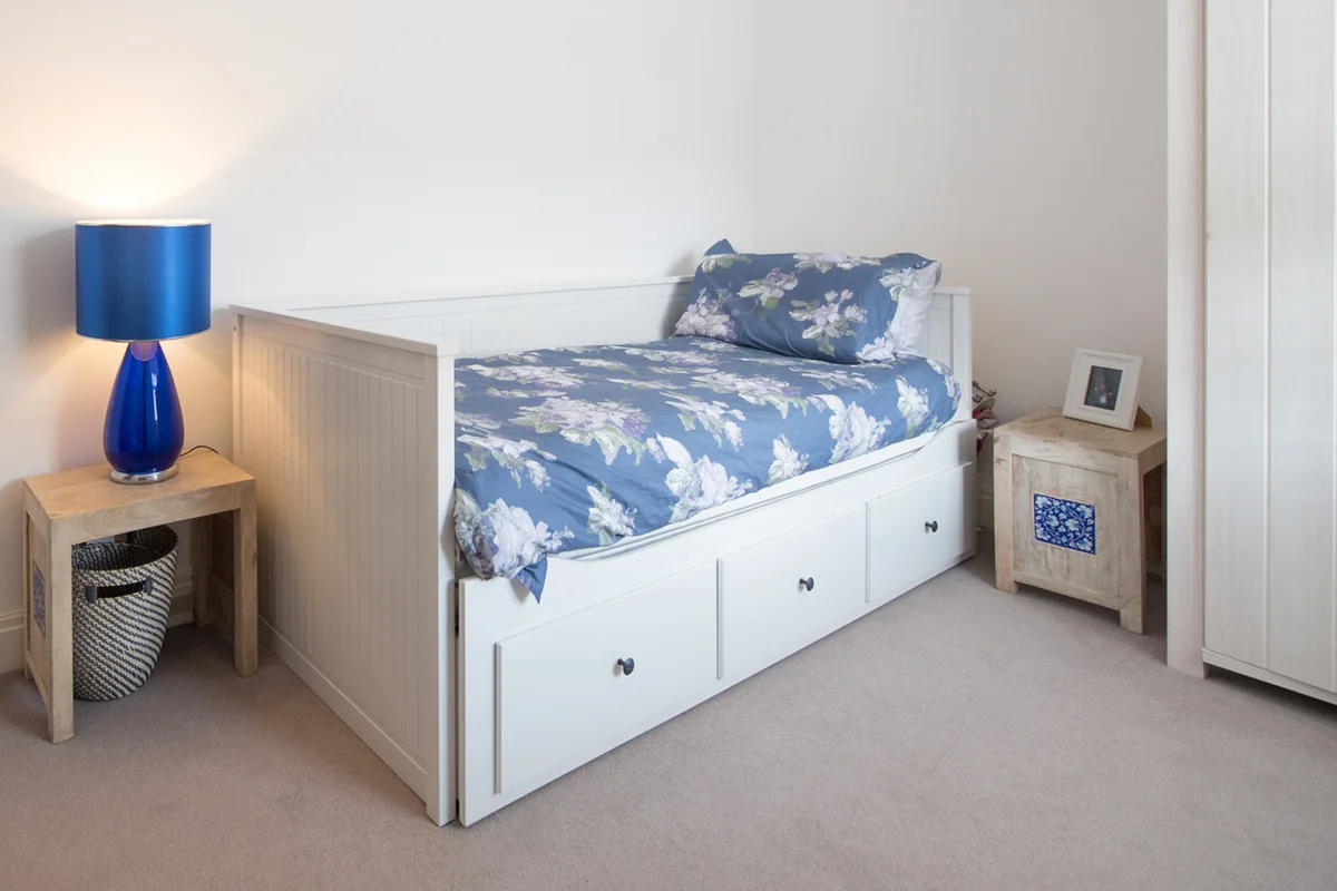A general view of a single bedroom with white walls and blue floral bedding with a blue coat hanging of the back of a door inside of a home