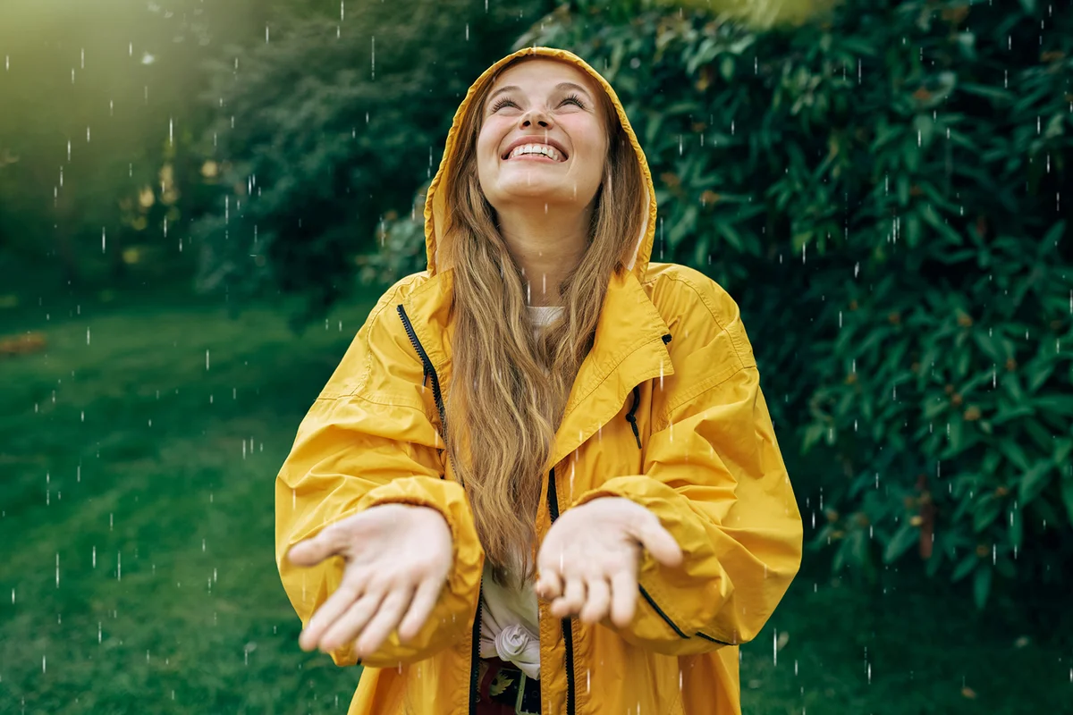 Image of a positive young blonde woman smiling wearing yellow raincoat during the rain in the park