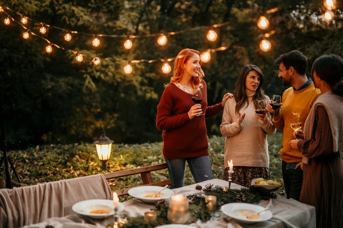 Group of elegantly dressed friends having a small boho dinner/wedding outdoors in a garden, drinking wine, and talking to each other with a great mood and smiles.