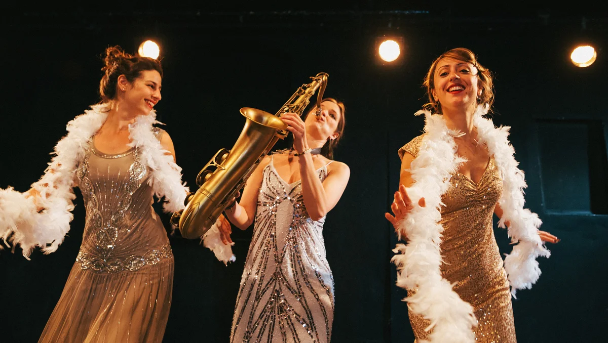 Three women wearing glittery flapper outfits dancing and playing instruments