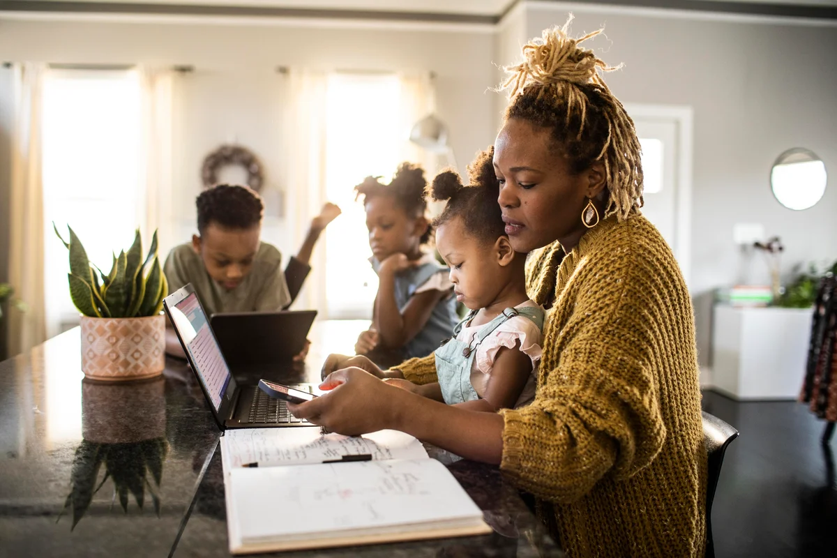 Mother working from home while holding toddler, family in background
