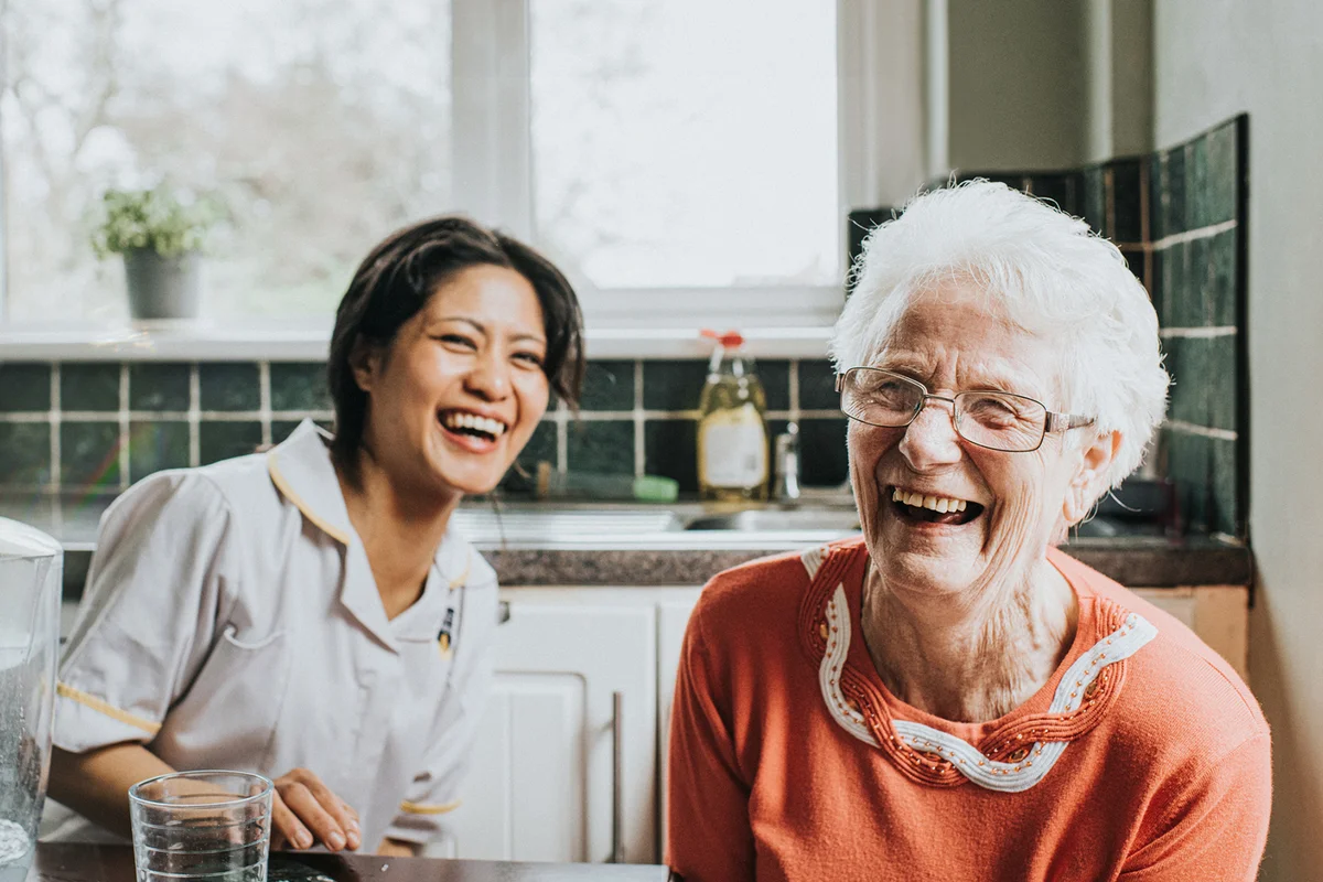 An elderly woman laughs beside a friendly young care assistant