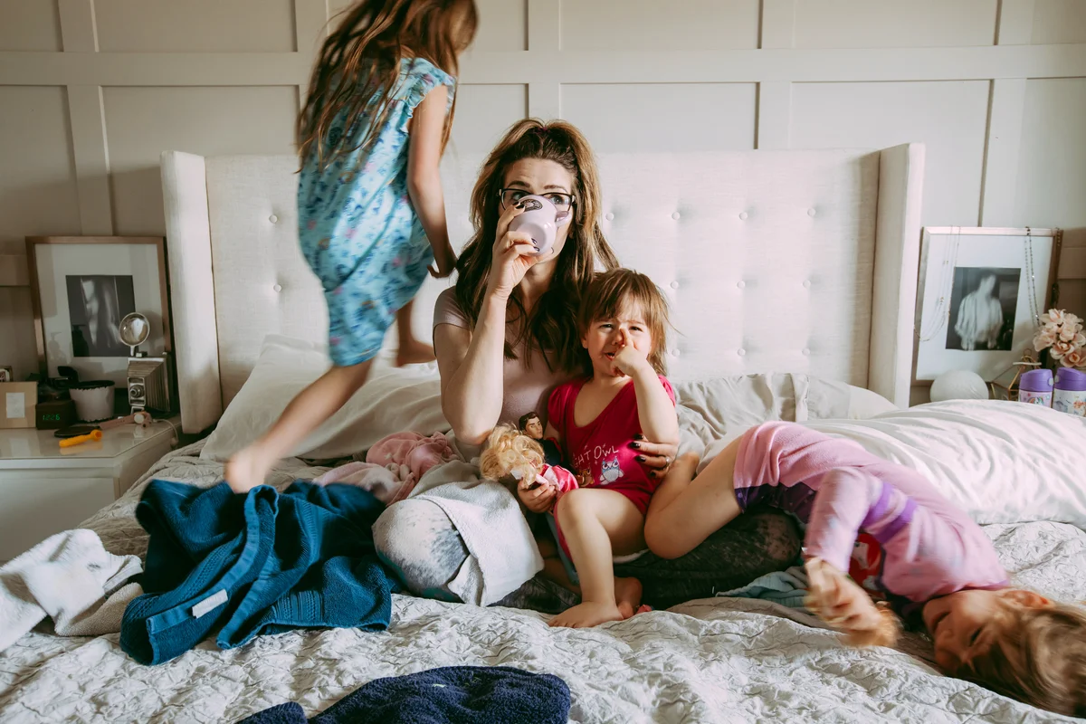 Young mother sipping on a cup of coffee on her messy bed while her three daughters are playing around her