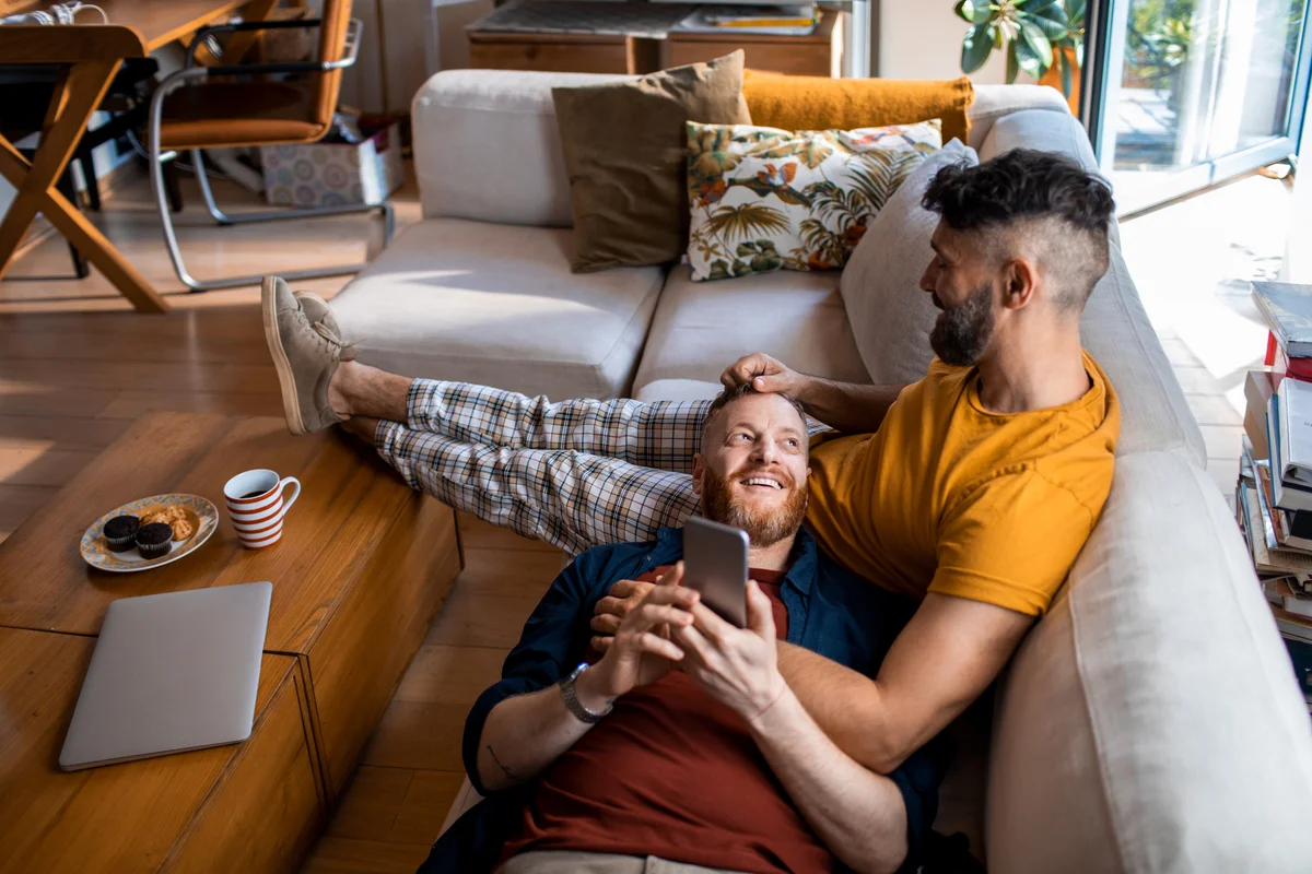 Male couple cuddling on couch in the morning looking at phone & talking