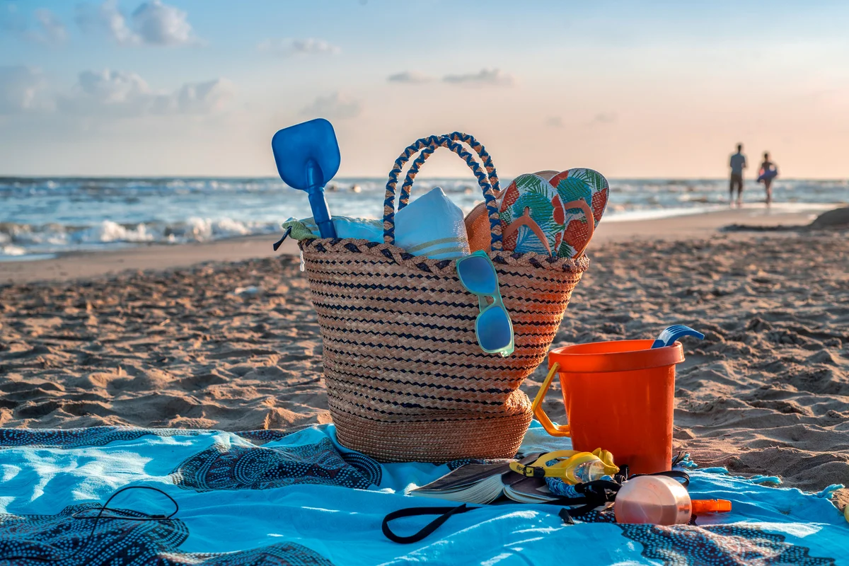 Beach bag with objects, glasses, towel, shovel, flip-flops, book etc that surface, laid on the beach at sunset