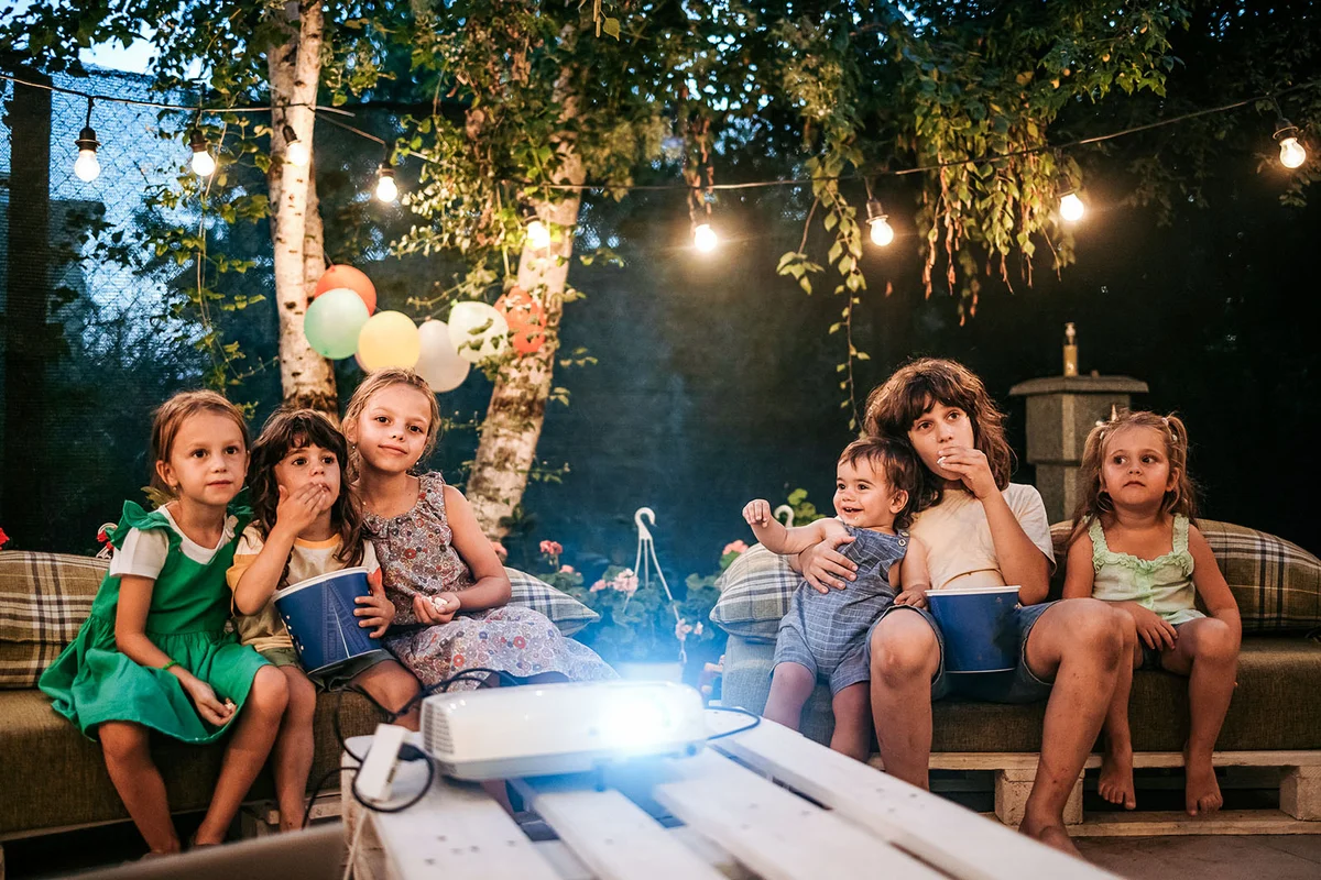 Group of children are sitting in the yard during a birthday party and watching a movie