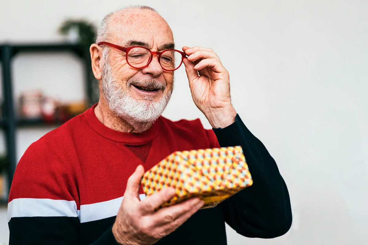 Smiling senior man wearing eyeglasses looking at gift box at home