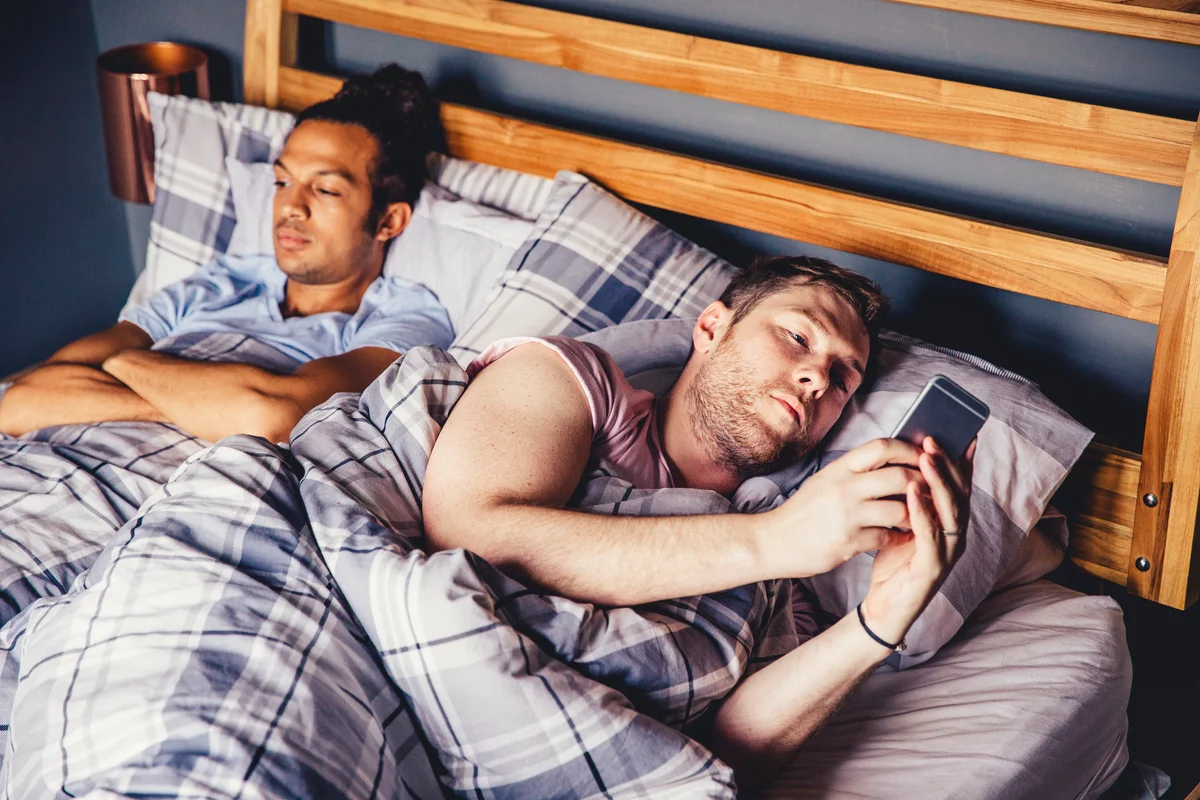 Male couple in bed, one with arms crossed unhappily while the other is rolled over on his phone.