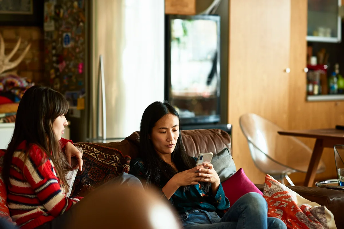 Women sitting on couch, one woman on her phone while the other tries to talk to her