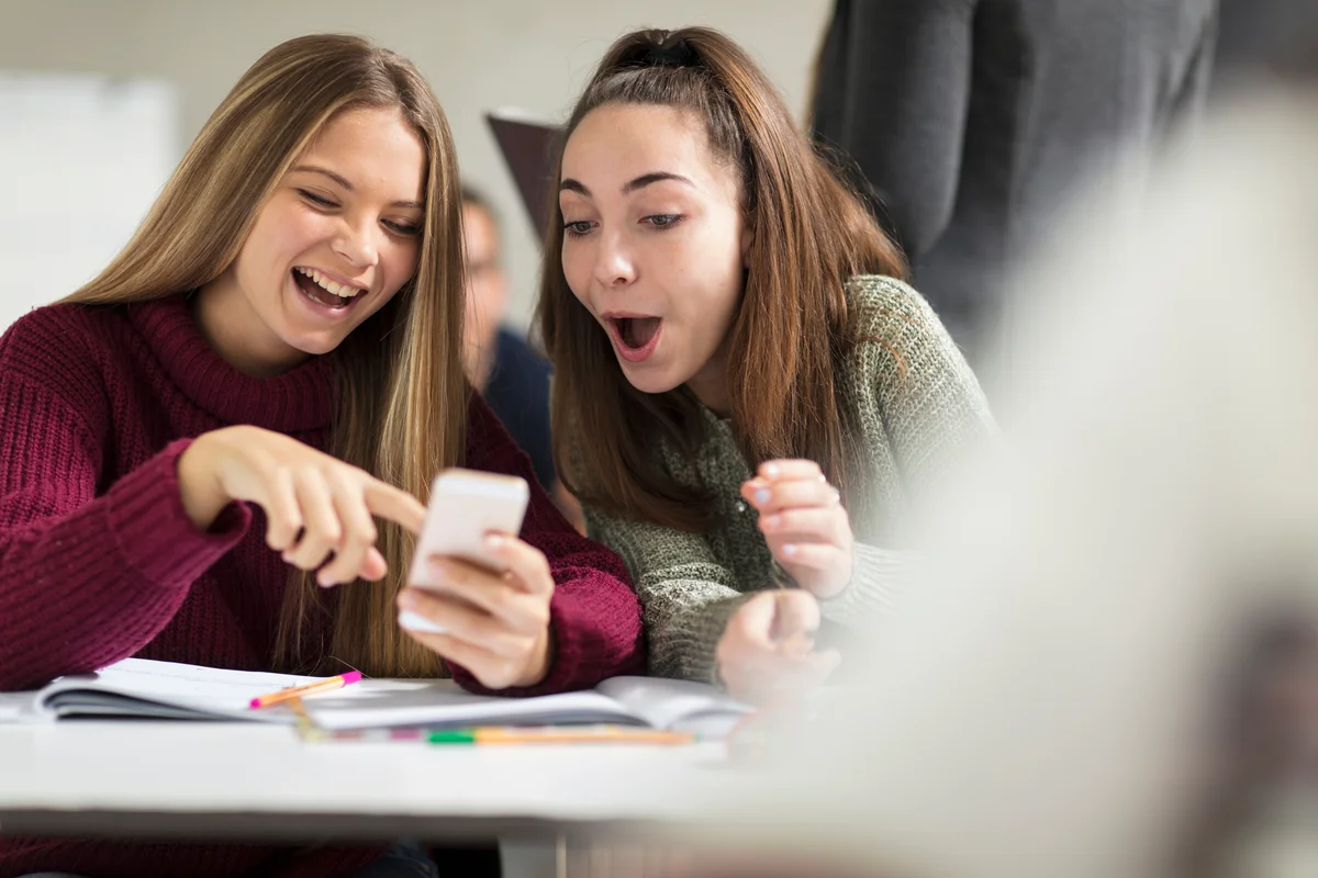 Teenage girls in class looking at cell phone and laughing about someone