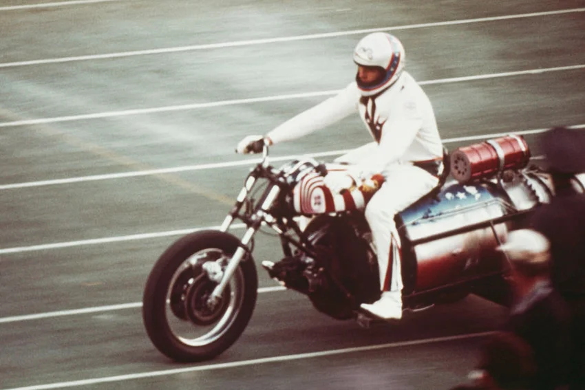 Evel Knievel rides a three wheeled motorcycle on an athletics track