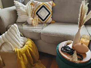 A living room sofa styled with a throw blanket and pillows and a tray full of decorative items. A basket of additional pillows is next to the sofa and a decorative ottoman is in front of the sofa with more decor on top. 