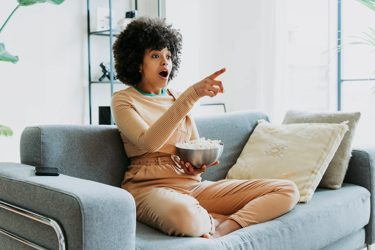 Shocked woman watching TV sitting on sofa with bowl of popcorn pointing at home