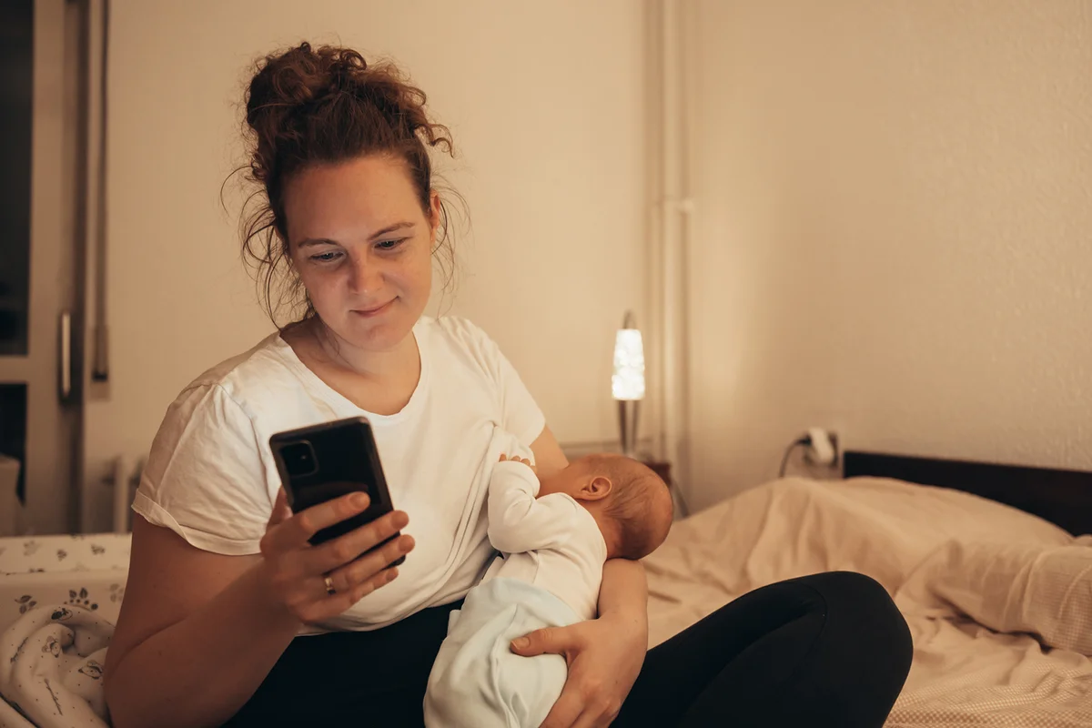 mother breastfeeding her newborn child at home while using a smartphone