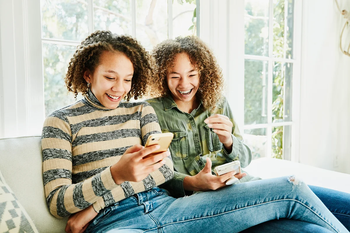 Laughing sisters sitting on couch in living room looking at smart phones