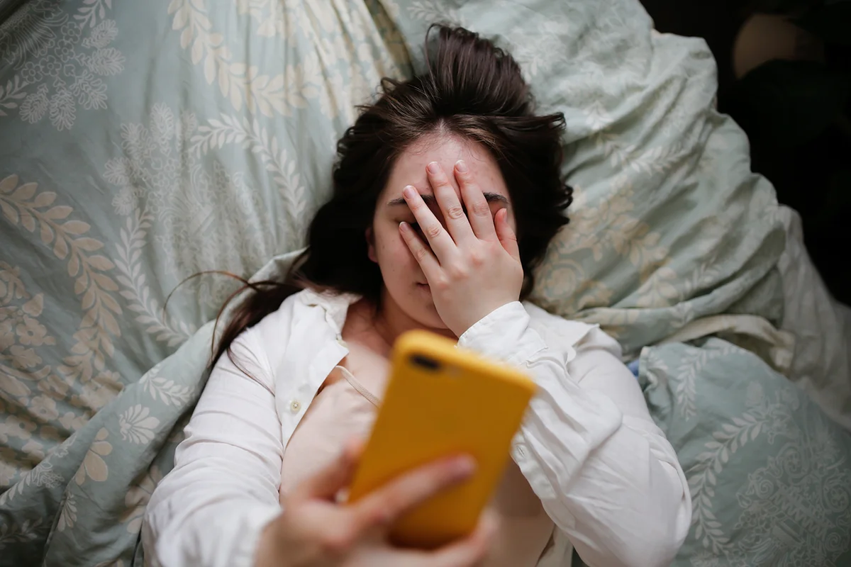 Young woman using phone on bed, close facepalm