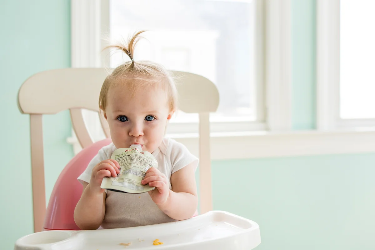 baby girl eating apple sauce from pack while sitting on high chair at home