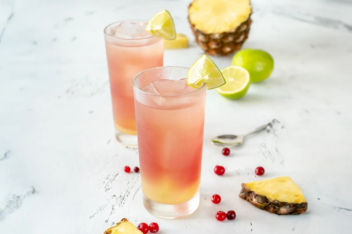 Two glasses of Bay Breeze cocktail garnished with lime wedges, served with pineapple slices and fresh cranberries