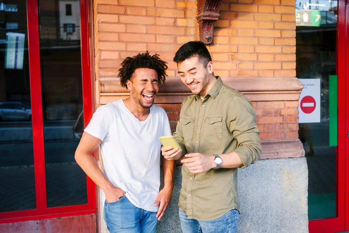 Young man laughing by friend using mobile phone near wall