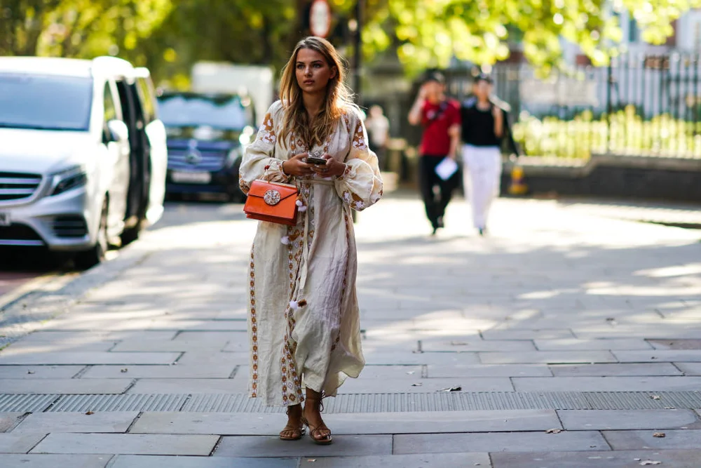 LONDON, ENGLAND - SEPTEMBER 15: A guest wears a white long dress with brown pixel geometric patterns, an orange leather bag, sandals, during London Fashion Week September 2019 on September 15, 2019 in London, England.