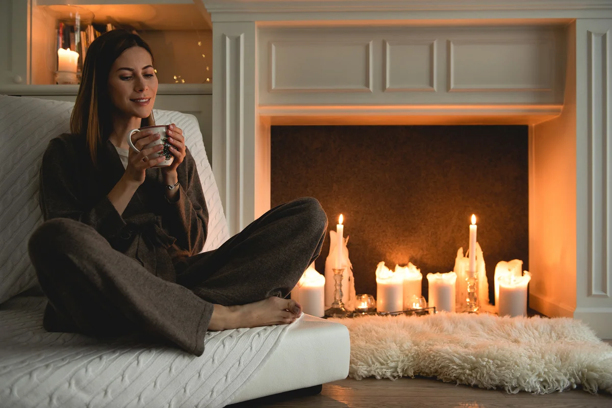 woman sitting cozy next to the fireplace with candles, holding a cup