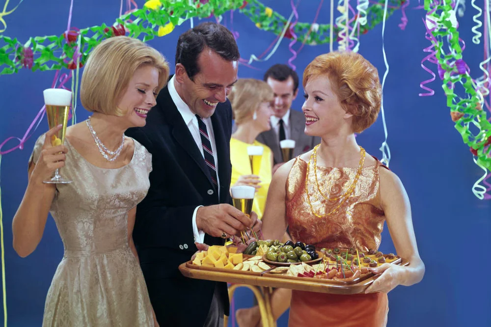 People at a 1950s party drinking alcohol and gathered around an hors d'oeuvre tray