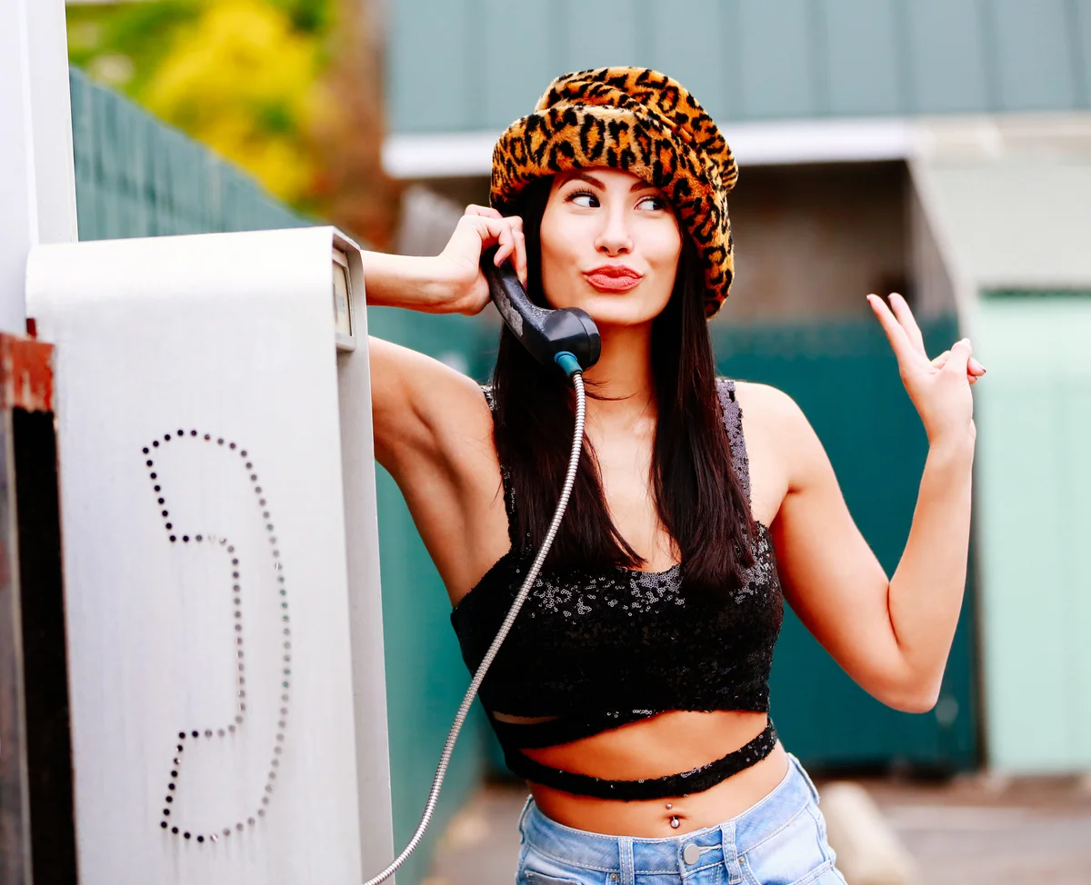 90s woman wearing cheetah hat talking on payphone and giving a peace sign