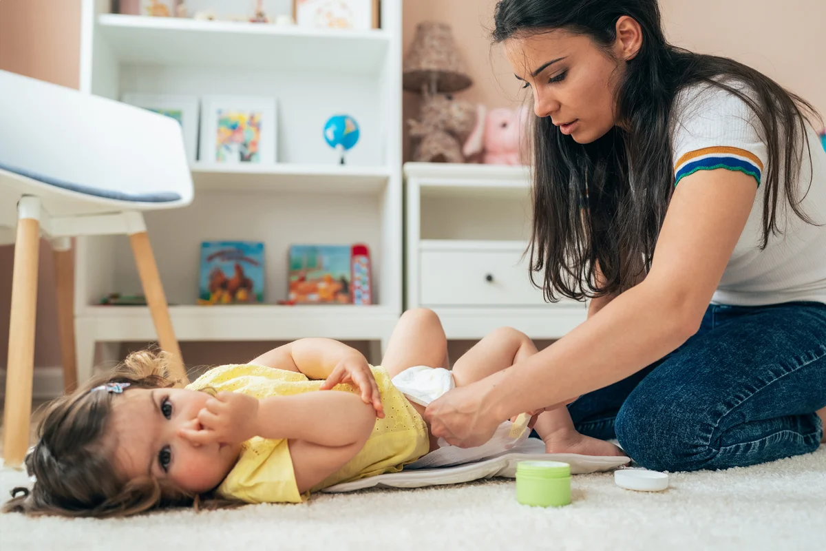 Mother changing diaper on toddler daughter on the floor at home