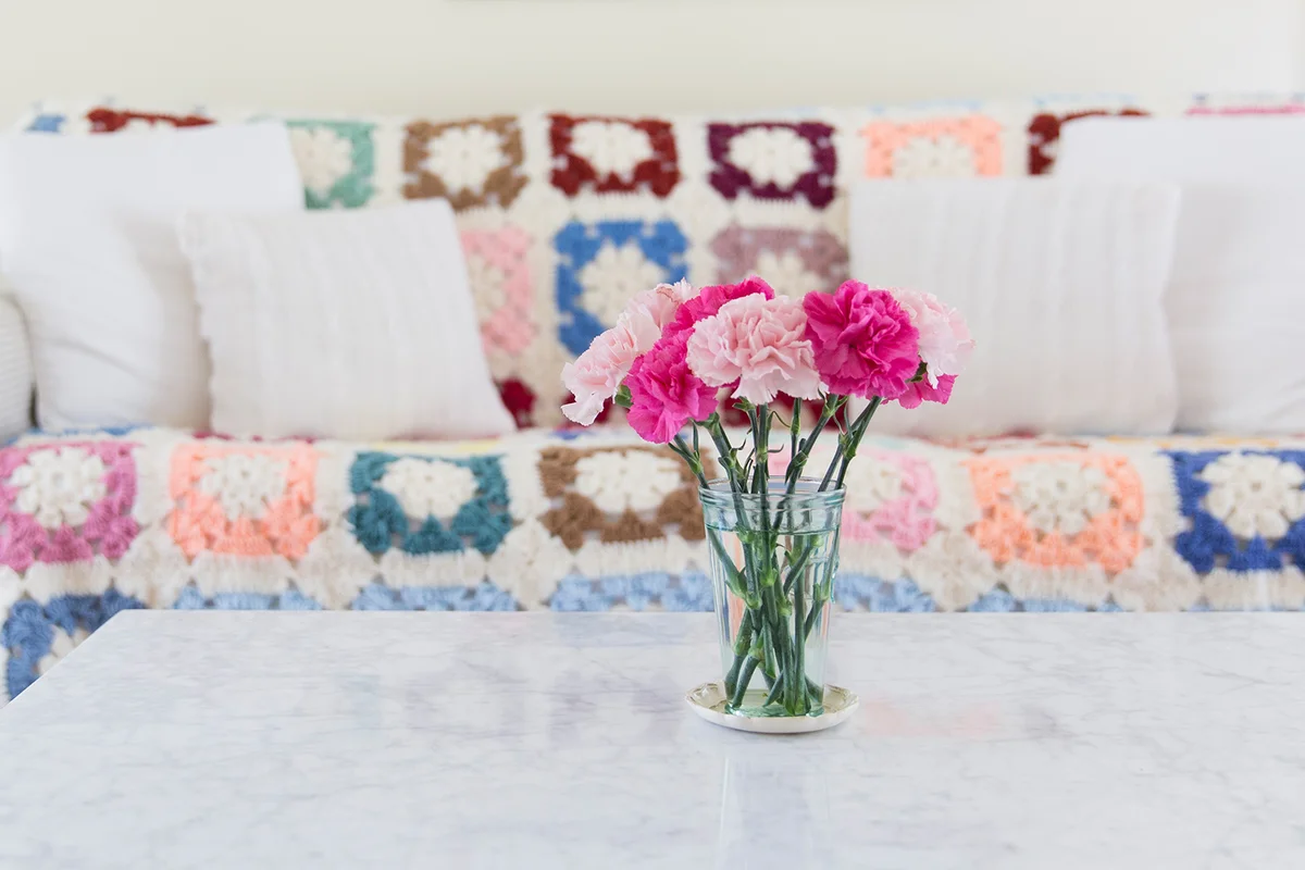 Pink carnations on marble coffee table in front of couch covered in a brightly colored granny square afghan
