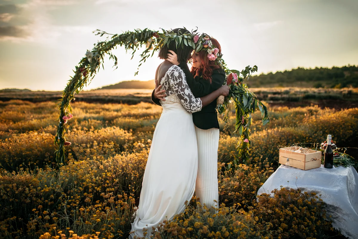 Two women standing under a boho wedding arbor covered in vines in a field