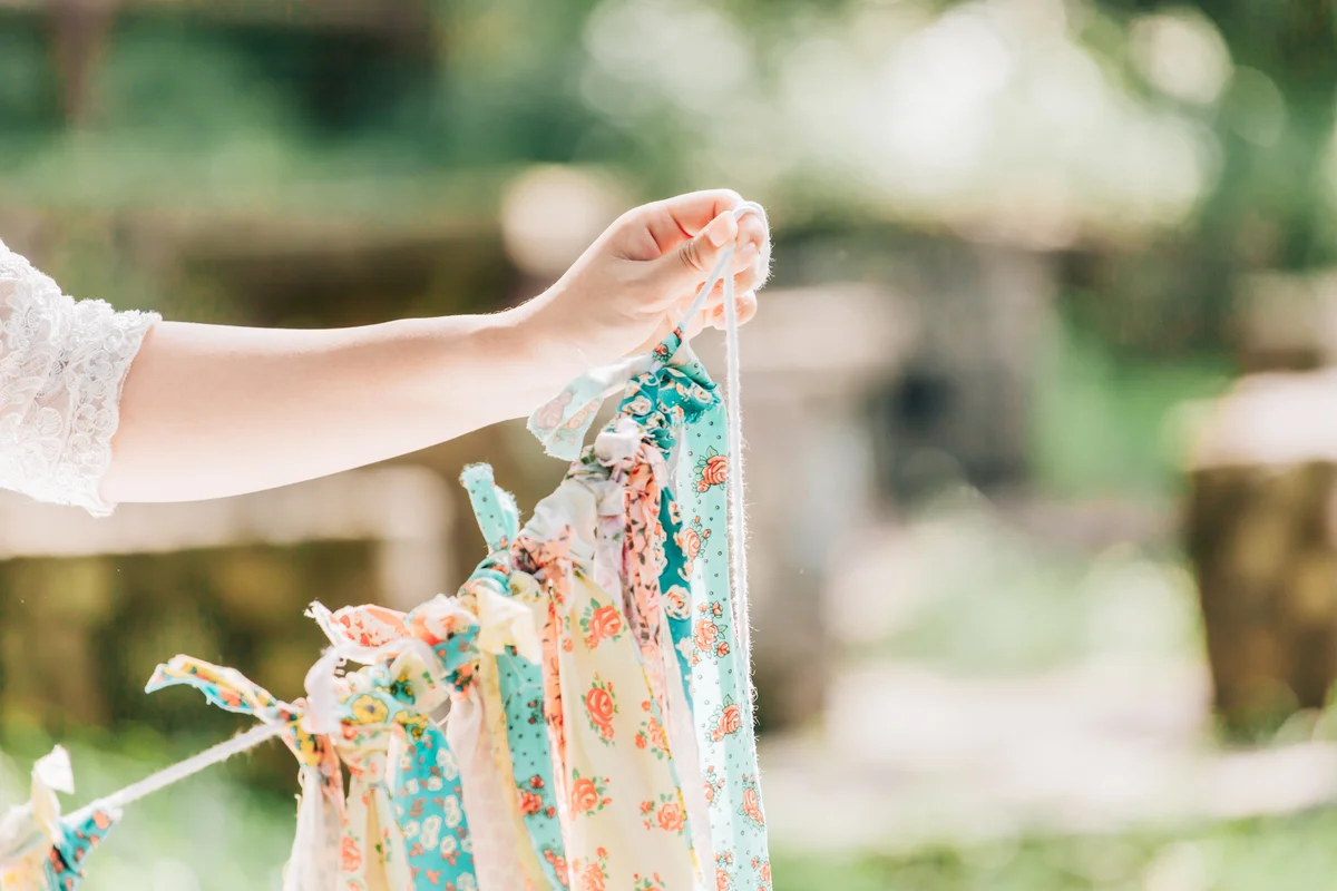 A bride is holding a row of colorful pennons for wedding party on a string against tree in a nature park.