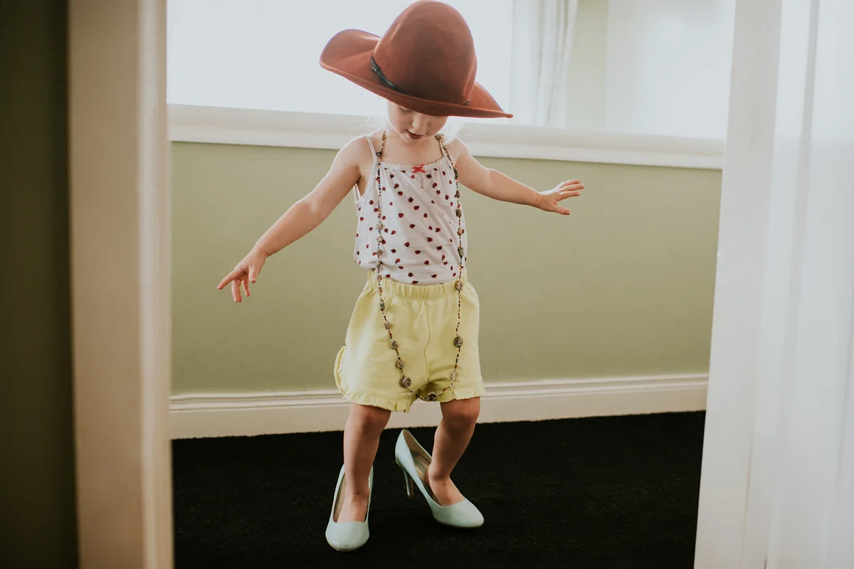 Cute toddler girl, dressed up in a hat, necklace and high heels.