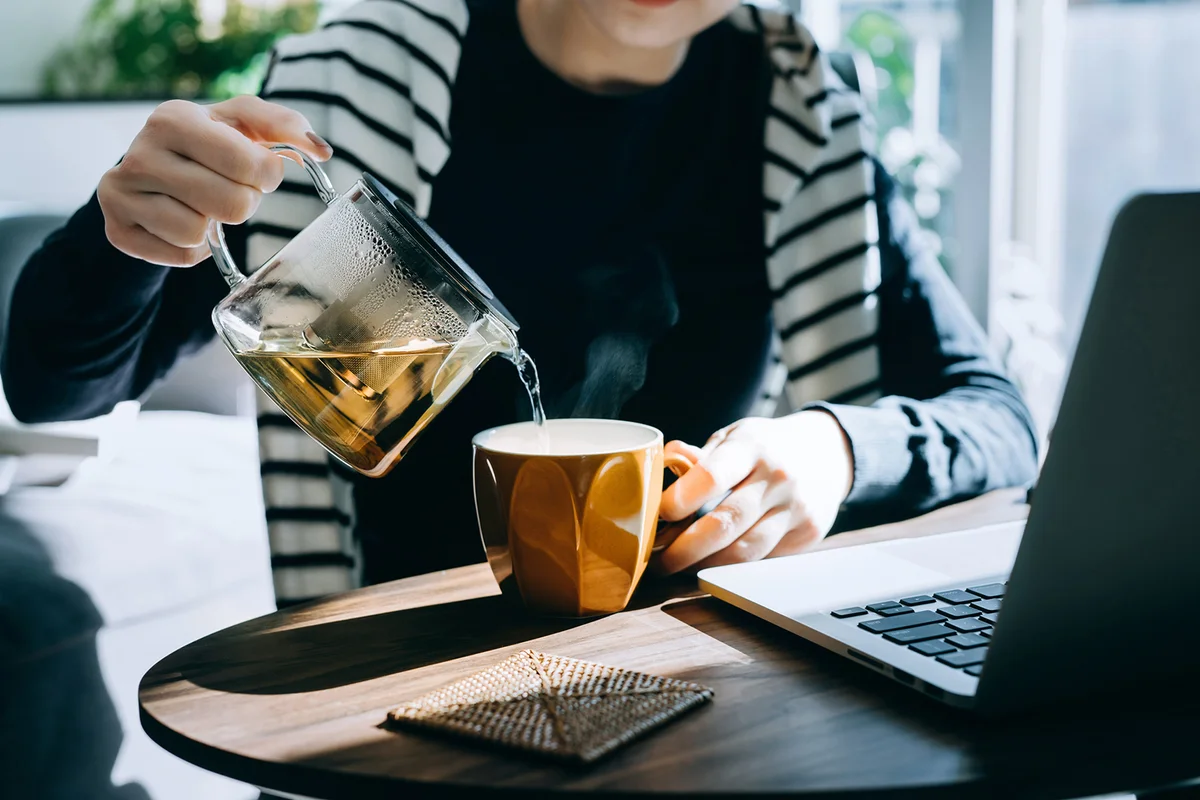 woman pouring a cup of tea from a stylish transparent tea pot into a cup